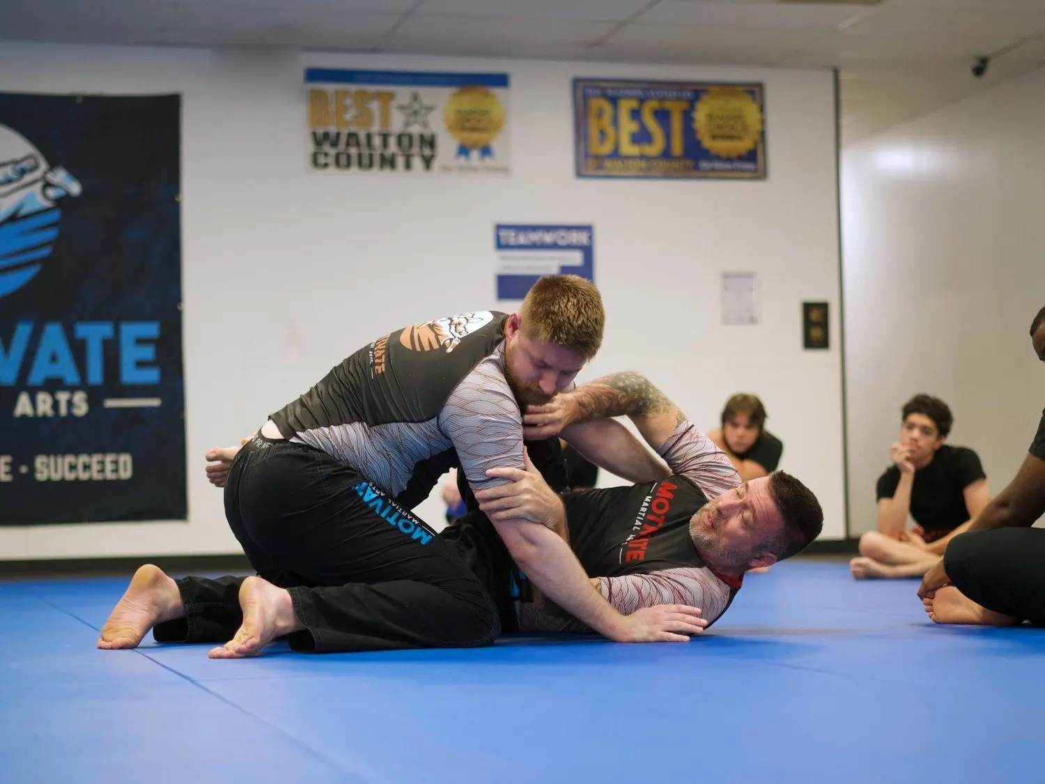 Two adults grappling No-Gi in rashguards on the blue mats in front of the Motivate banner while students watch