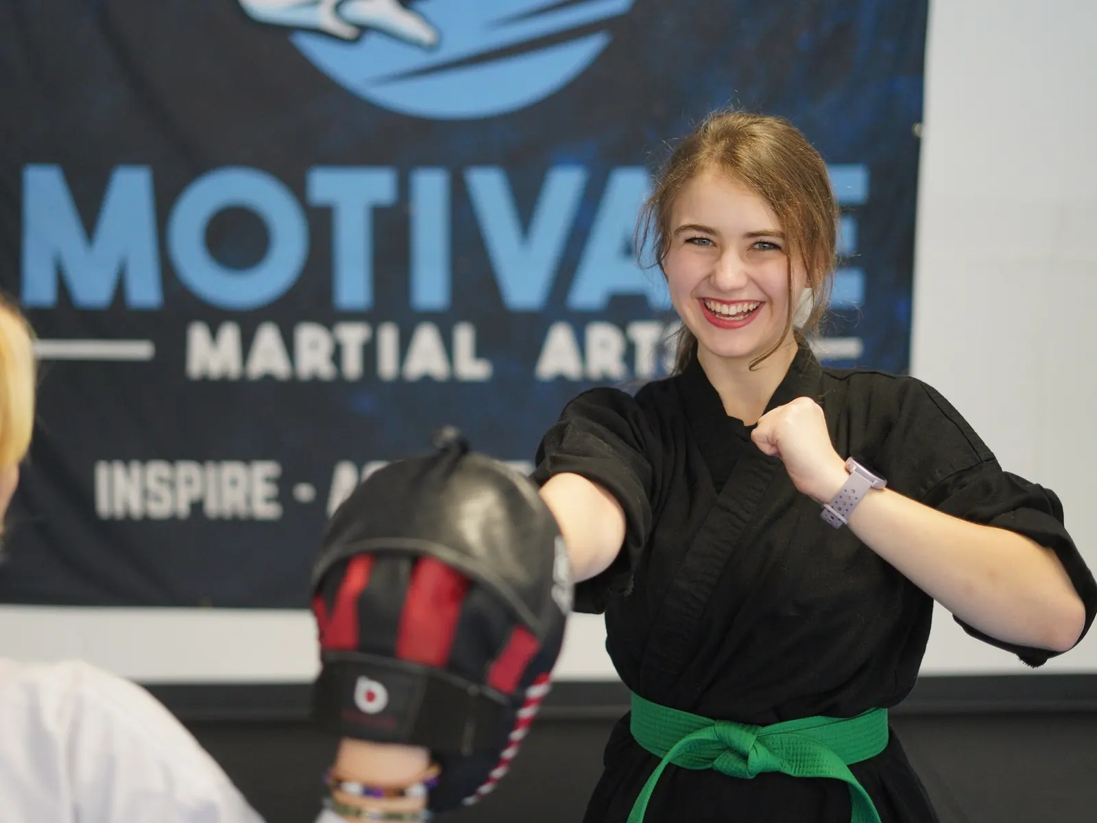 Smiling TaeKwonDo student in a green belt punching a focus mitt in front of the Motivate banner